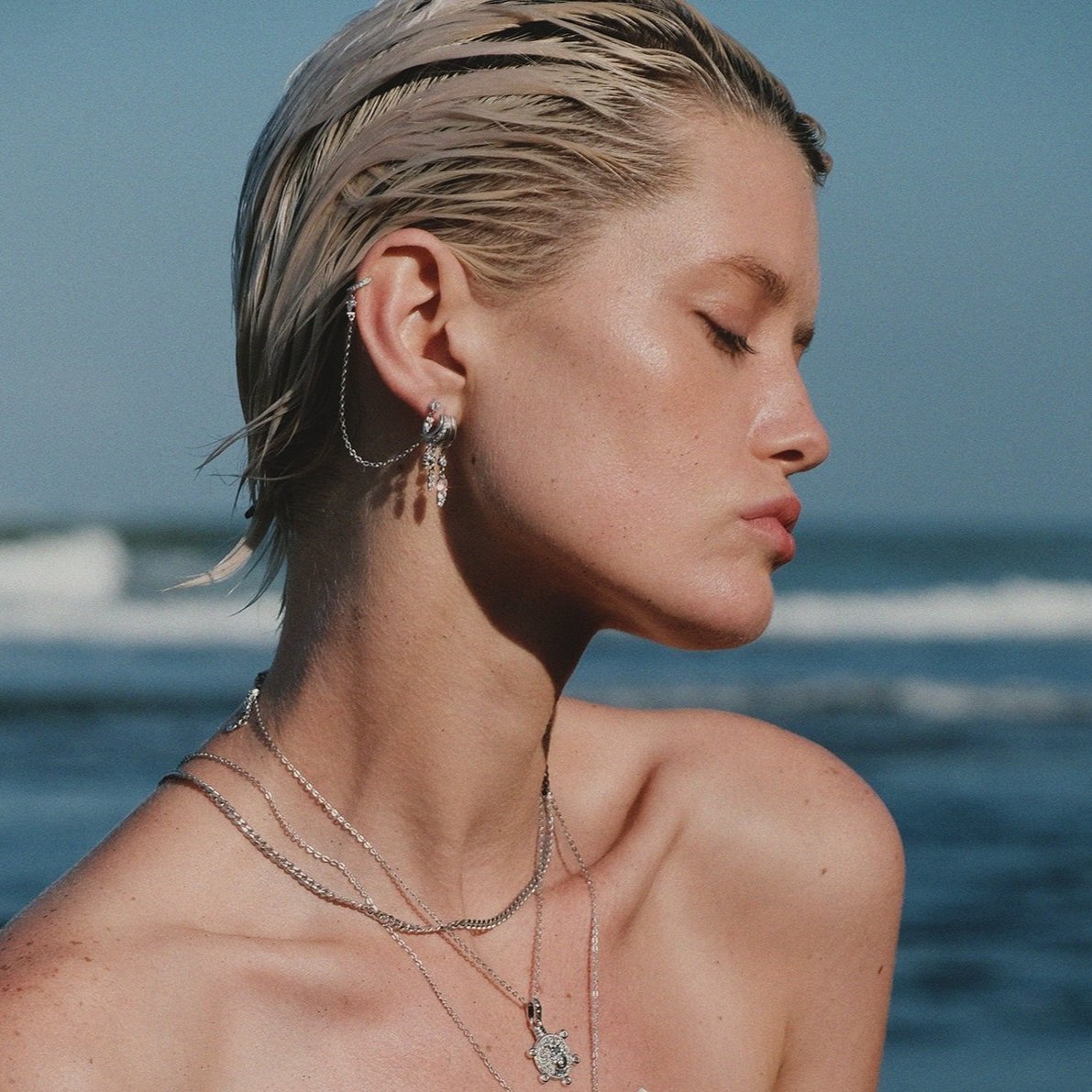 Model wearing layered silver necklaces and ear cuffs at beach.