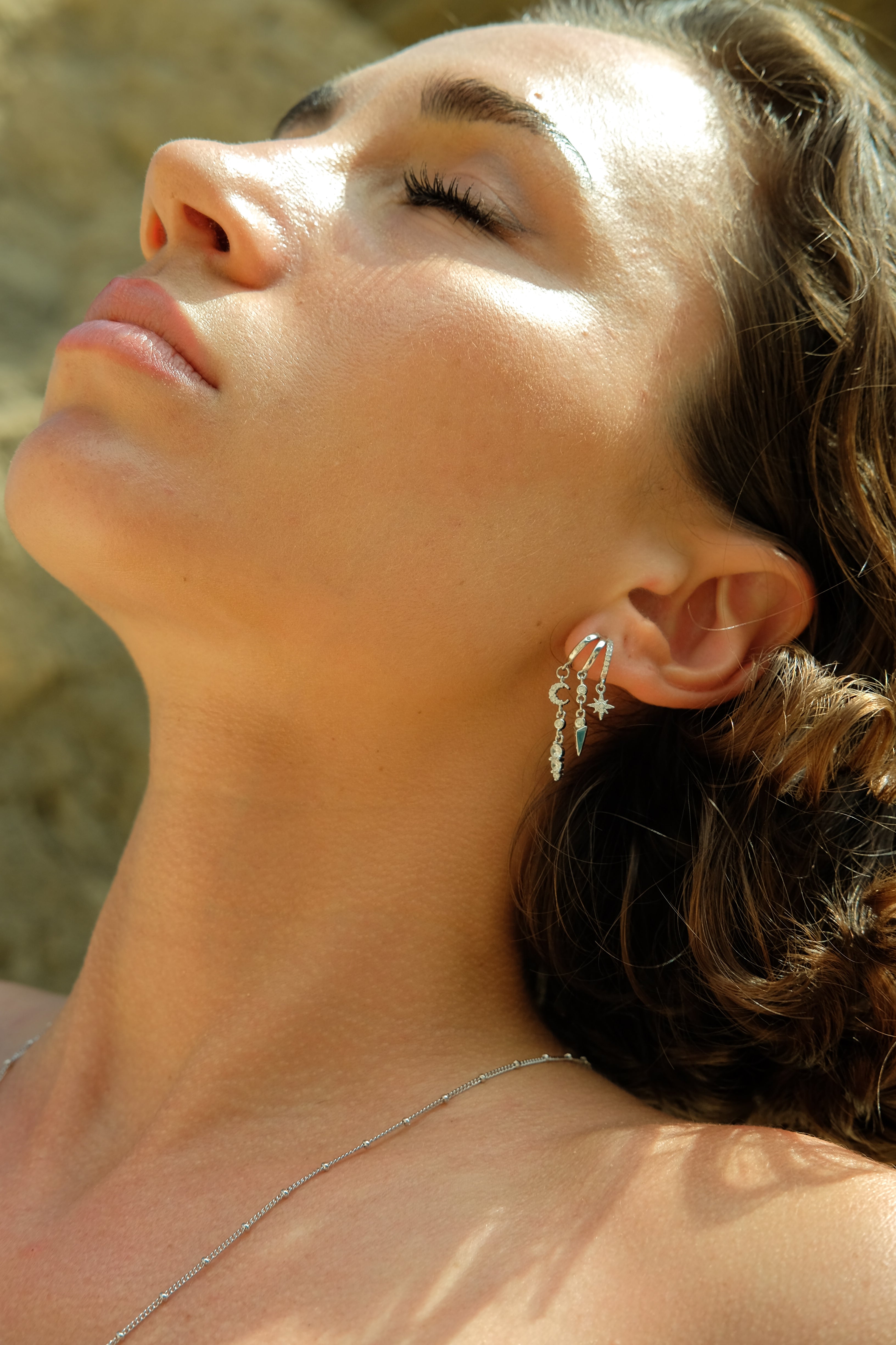Woman wearing silver drop earrings with layered chains, basking in sunlight. Elegant jewelry from Aaria London.