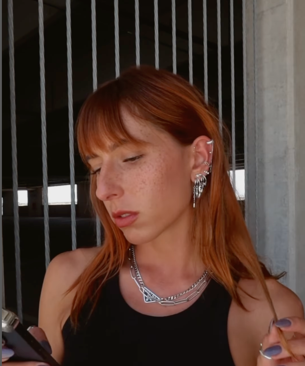Stylish woman with red hair wearing silver angel hoop earrings and a chain necklace, holding a vape.