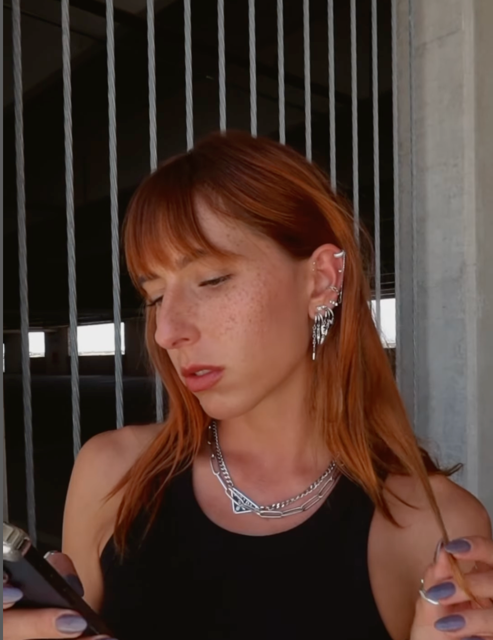 Stylish woman with red hair wearing silver angel hoop earrings and a chain necklace, holding a vape.