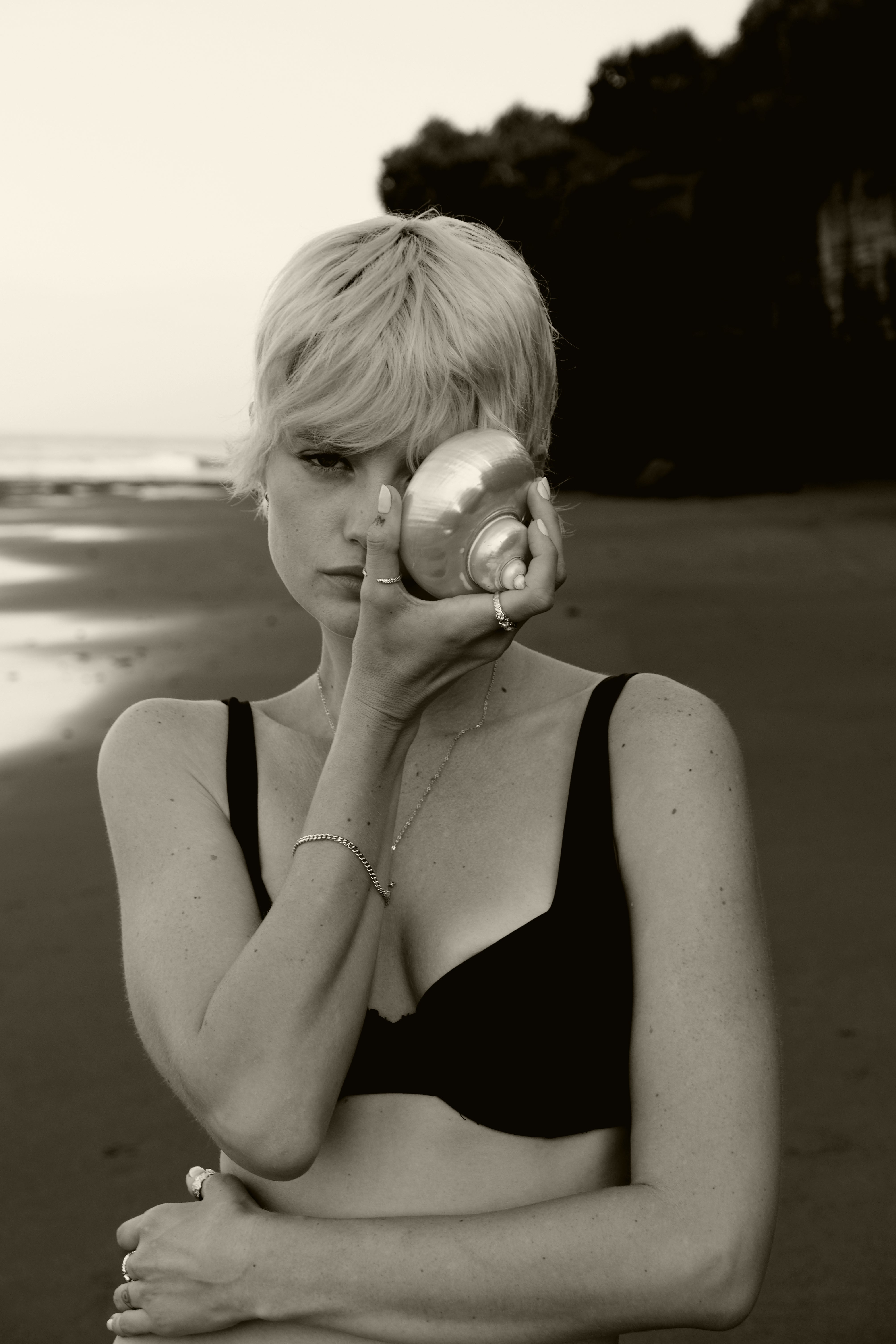 A woman with short blonde hair in a black bikini top holding a seashell to her eye on a sandy beach, in sepia tone.
