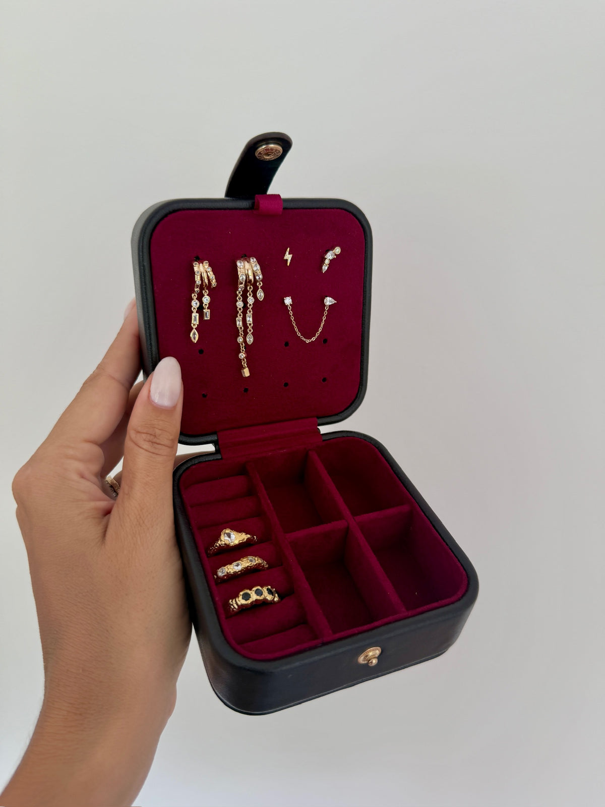 Black jewelry box with red interior holding gold earrings and rings, held by a hand against a plain background.