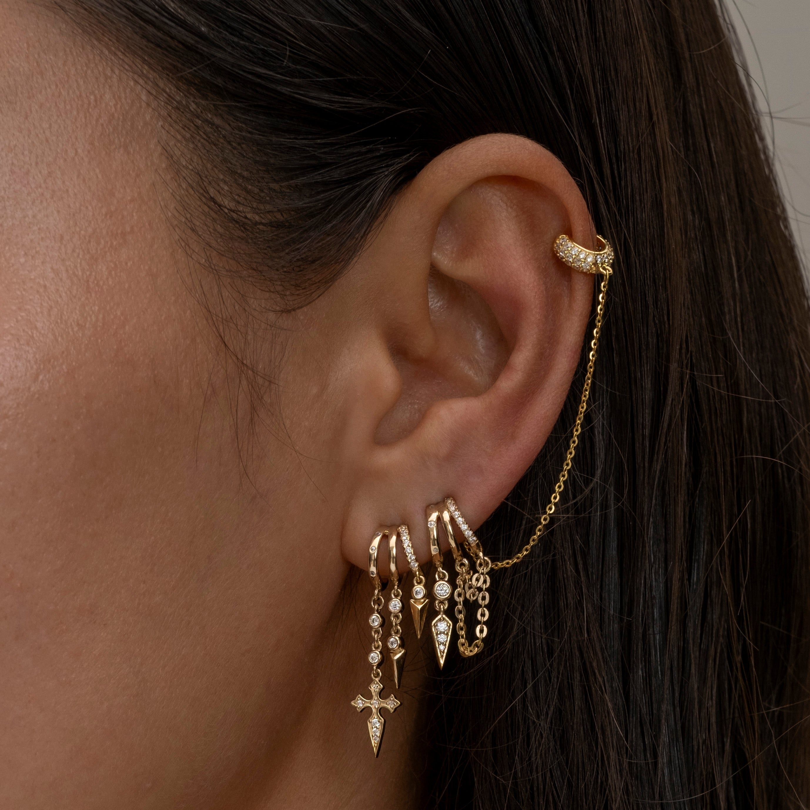 Close-up of an ear with gold earrings and a ring, against a neutral background.
