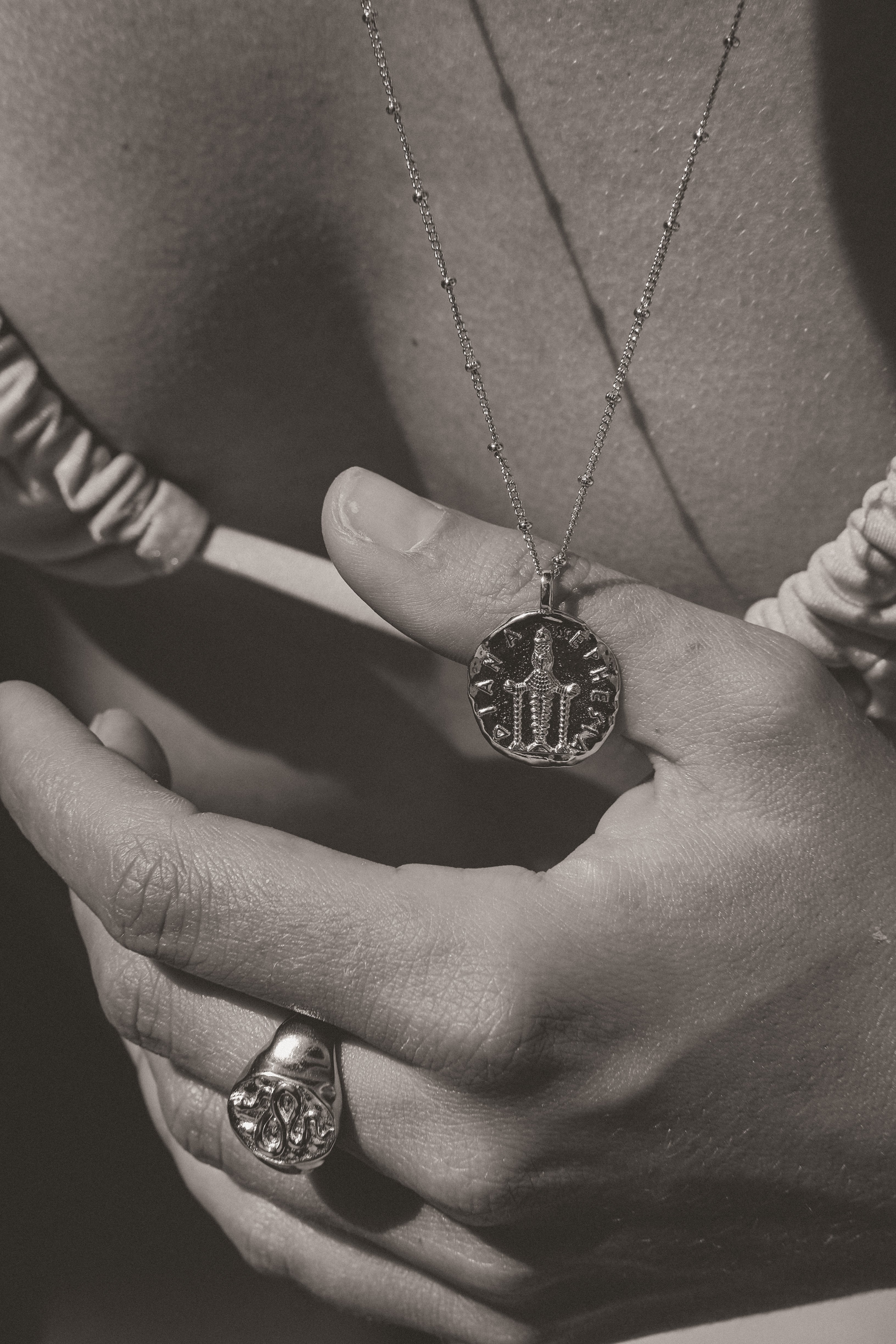 Aaria London engraved coin pendant necklace and silver ring worn on a hand, showcasing timeless craftsmanship.